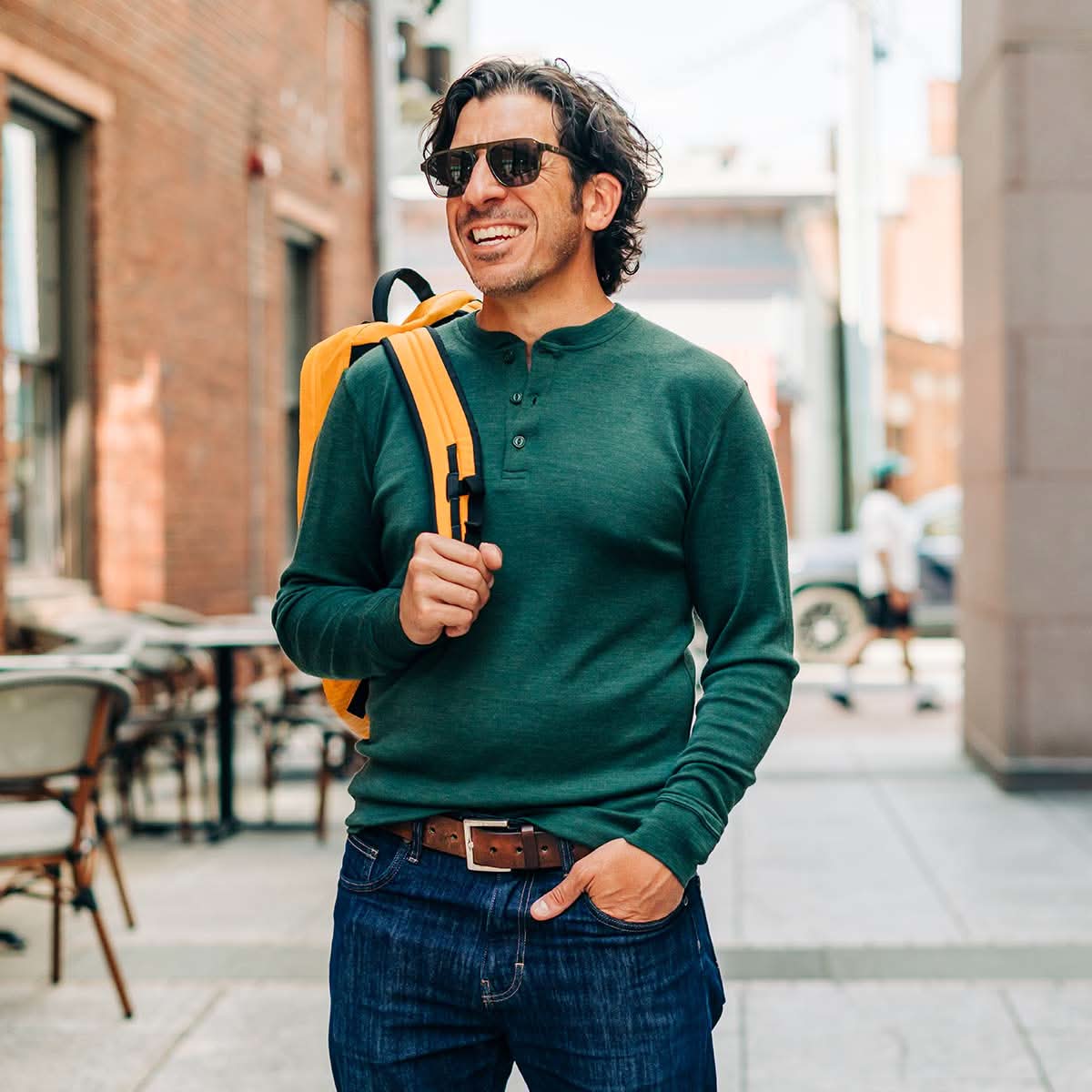 Man wearing a green merino wool sweater and sunglasses, holding a yellow backpack in an urban setting.