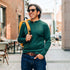 Man wearing a green merino wool sweater and sunglasses, holding a yellow backpack in an urban setting.