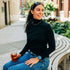 Woman sitting on a bench holding a drink, wearing a black merino wool turtleneck sweater and blue jeans, with a blurred outdoor background.