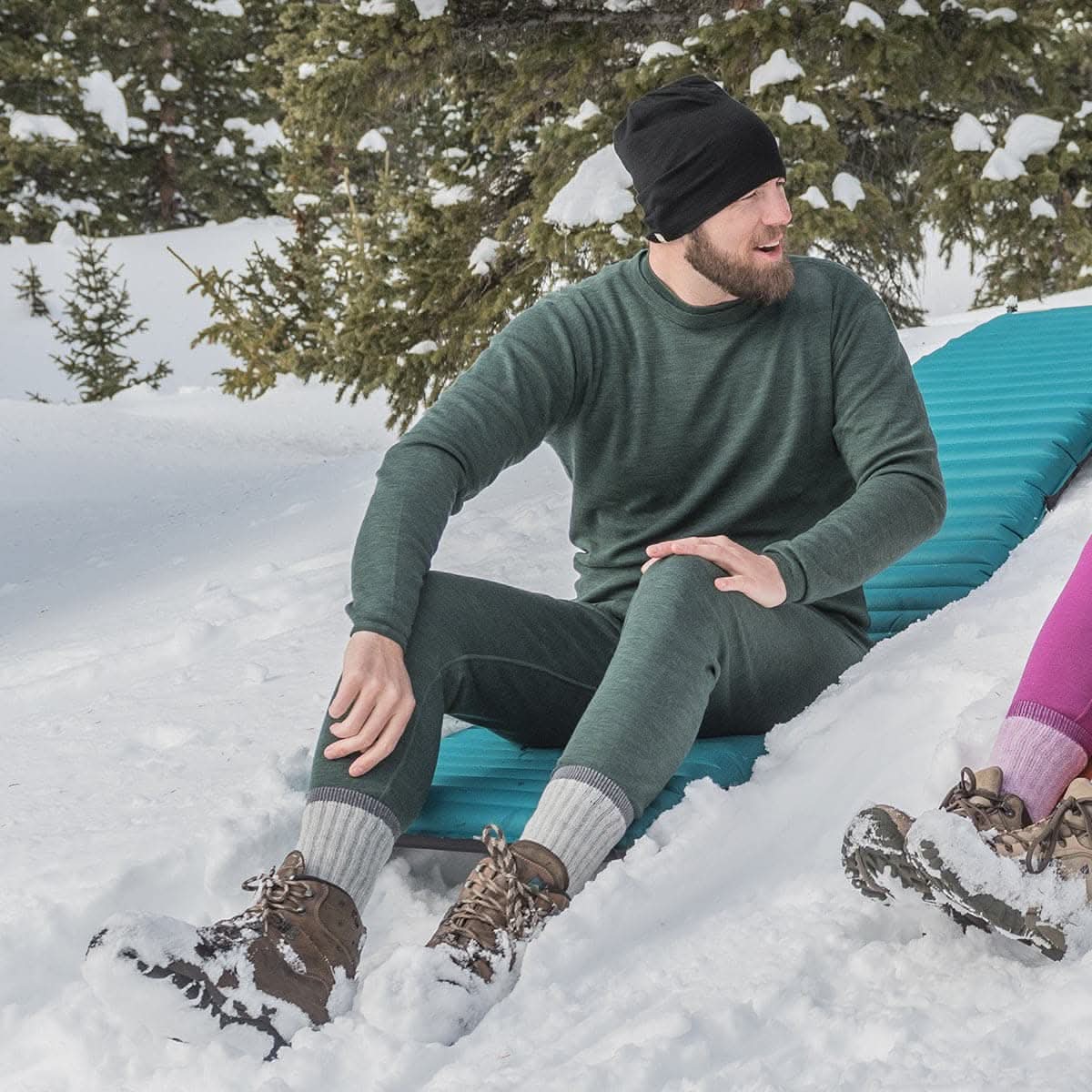 Minus33 Merino Wool Clothing Kancamagus Men's Midweight Wool Bottom thermal baselayer, man seated on a sleeping pad in snow wearing forest-green base layers