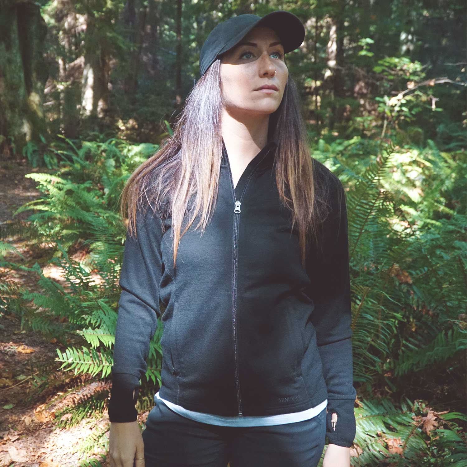 A woman stands in front of tall ferns surrounded by autumn leaves on the ground. Sunlight peeks through the trees. The woman is wearing a hundred percent merino wool heavyweight full zip sweater. This outer layer features a two and a half inch mock neck collar, two zippered pockets, and thumbholes.