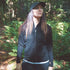A woman stands in front of tall ferns surrounded by autumn leaves on the ground. Sunlight peeks through the trees. The woman is wearing a hundred percent merino wool heavyweight full zip sweater. This outer layer features a two and a half inch mock neck collar, two zippered pockets, and thumbholes.
