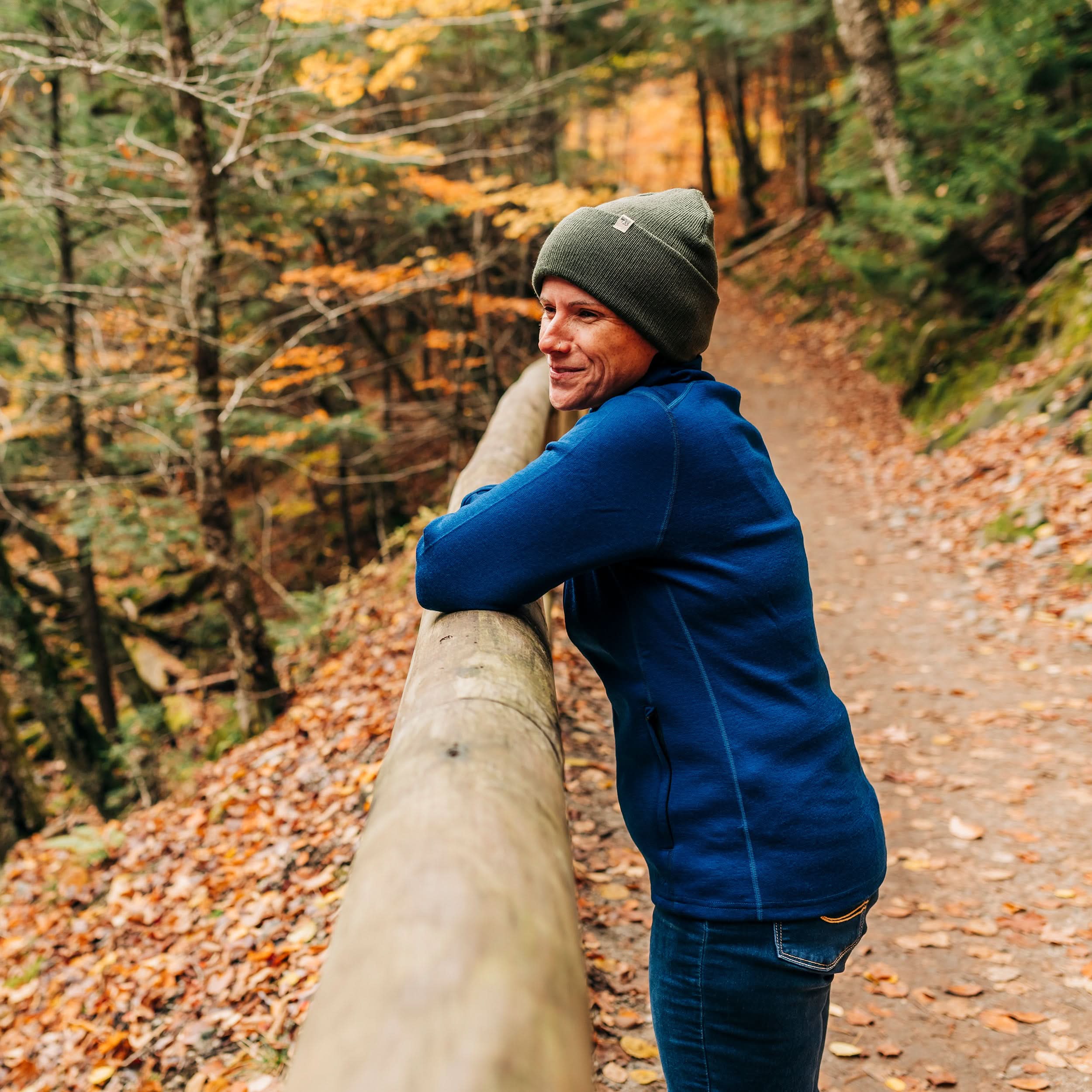 A woman stands on a walking trail in autumn, wearing her hundred percent merino wool heavyweight full zip sweater and a hat. This outer layer features a two and a half inch mock neck collar, two zippered pockets, and thumbholes.
