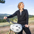 Front three-quarter action view of a woman cycling in a black micro weight merino wool quarter-zip top, showing the stand collar, partial zipper, fitted sleeves, shaped torso, and lightweight fabric