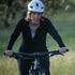 Wide side action view of a woman cycling in a black micro weight merino wool quarter-zip top, showing the raised collar, fitted long sleeves, curved riding posture, and lightweight fabric
