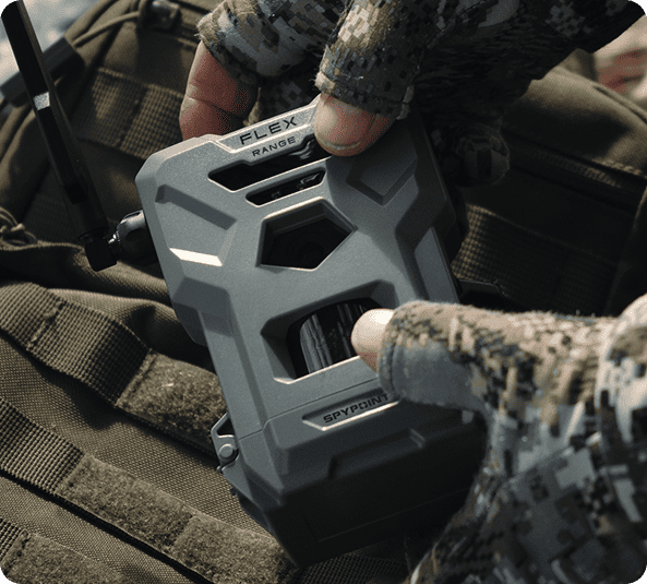 SPYPOINT FLEX-RANGE trail camera held in gloved hands, showing its front housing and antenna