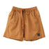 A pair of shorts in a warm camel brown color, featuring an elastic waistband with a drawstring, side pockets, and a small circular logo patch on the lower left leg, displayed against a solid white background.