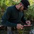 Three-quarter side view of a man wearing a forest green midweight merino wool long-sleeve henley, black cap, and sunglasses while preparing fishing gear beside a wooded stream
