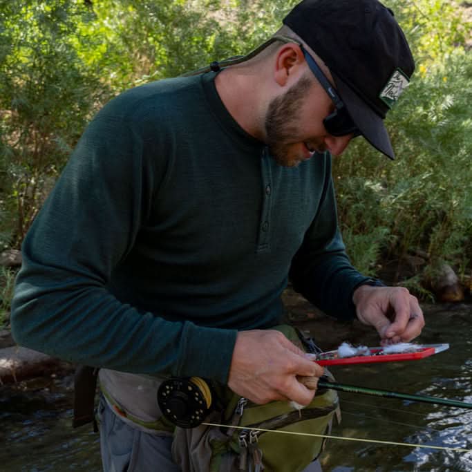 Angled front-side view of a man wearing a navy midweight merino wool long-sleeve henley with a black cap and sunglasses, looking down while arranging a fishing lure beside a shaded stream